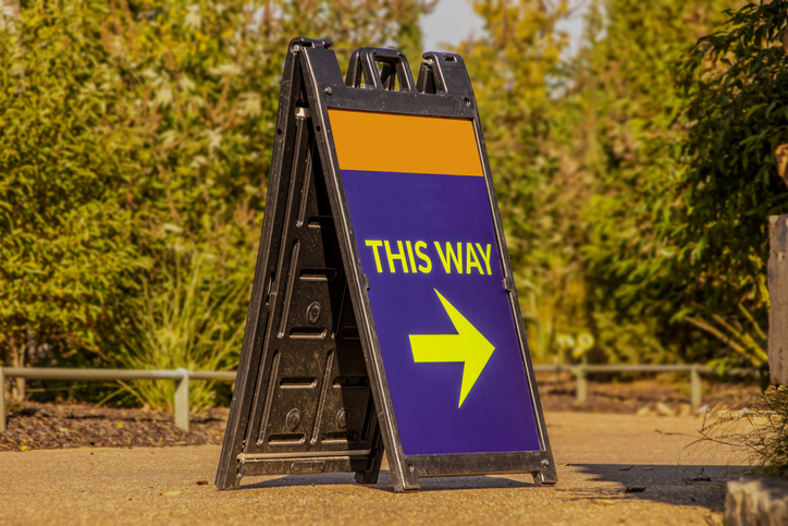 Room for Copy on Large blue and orange standing plastic sandwich sign sitting on paved trail with blurred foliage in background with arrow saying This Way